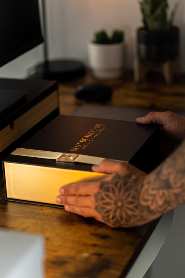 A person holding a box containing the Tobacco & Vanilla Gift Set Beard Grooming Kit with beard wax, surrounded by a potted plant and a book.