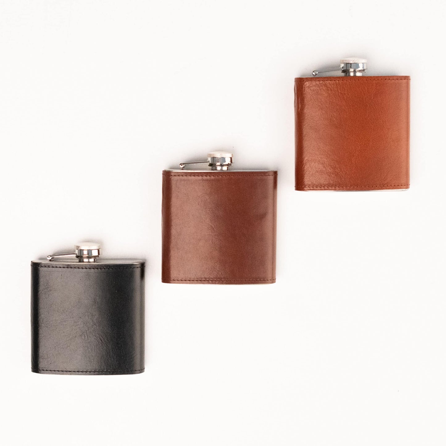 Hip Leather-Covered Flask: A group of stylish flasks, including a brown leather flask with a metal flask and a black flask with a silver top. Elevate your drinking experience with this European-made work of art, crafted from stainless steel and luxurious natural leather. Compact and sophisticated, it holds 177ml of spirits. Perfect for any occasion, fill it with your preferred libation and stand out from the crowd. From Men In Style, your destination for exclusive men's accessories.