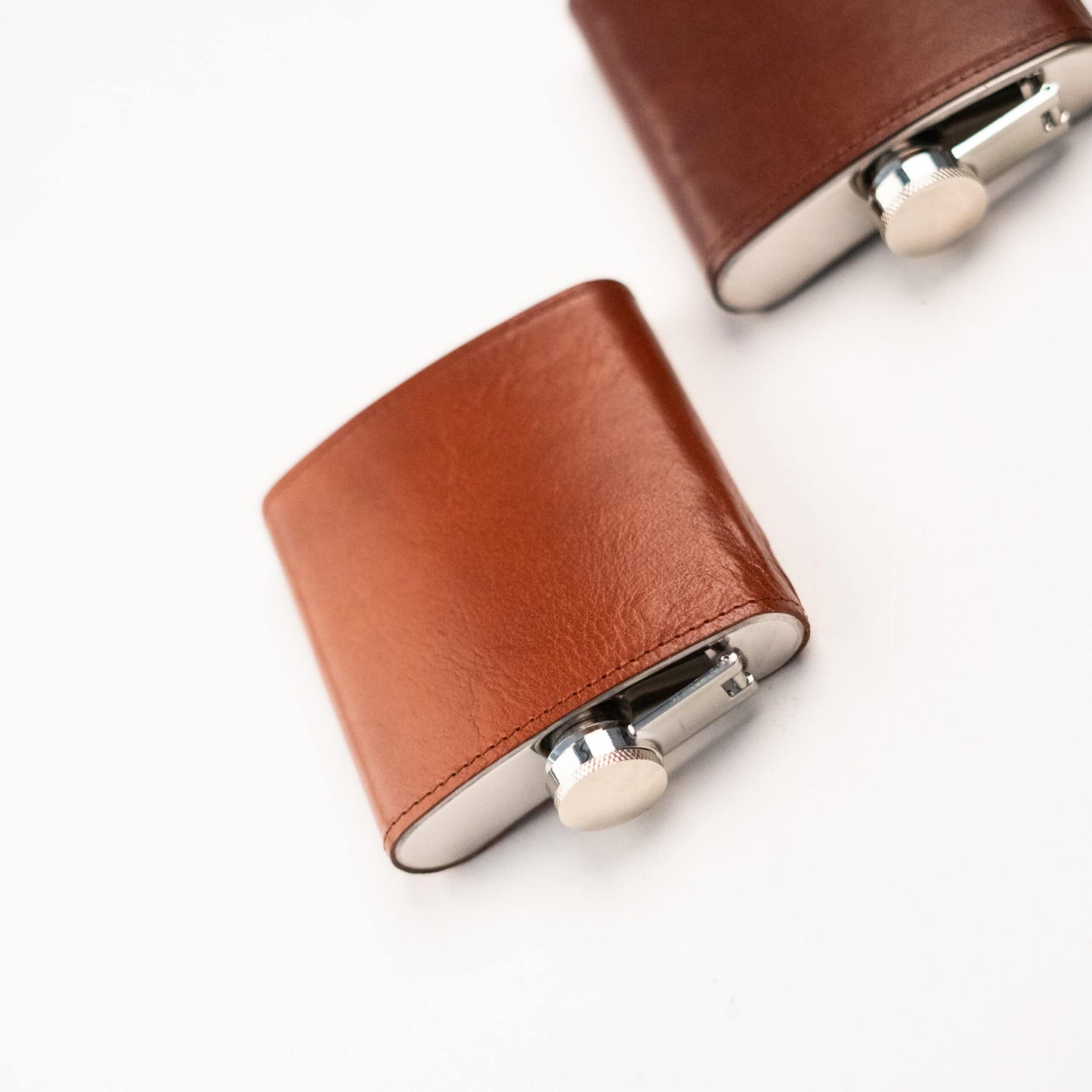 A pair of brown leather flasks, crafted from stainless steel and enveloped in luxurious natural leather. Perfect for sipping spirits or sharing a drink with friends. Elevate your drinking experience with the Hip Leather-Covered Flask from Men In Style.