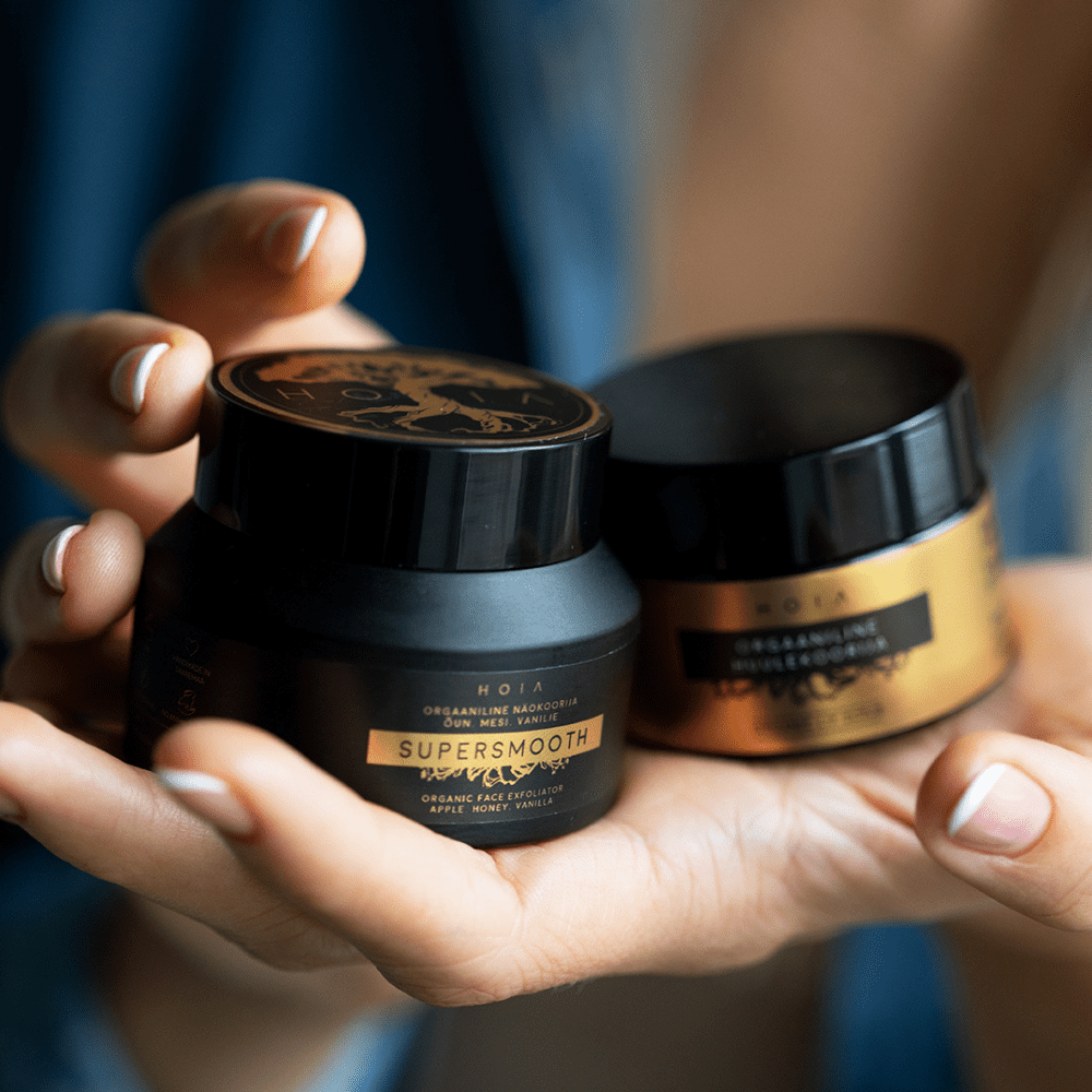 A person holding two jars of Organic Face Scrub, a natural exfoliator with apple water, olive stone powder, aloe extract, and vanilla pod.