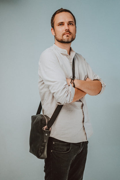 A man with crossed arms wearing the Small Shoulder Bag - Black, showcasing its genuine leather craftsmanship, adjustable shoulder strap, and multiple compartments for organization.