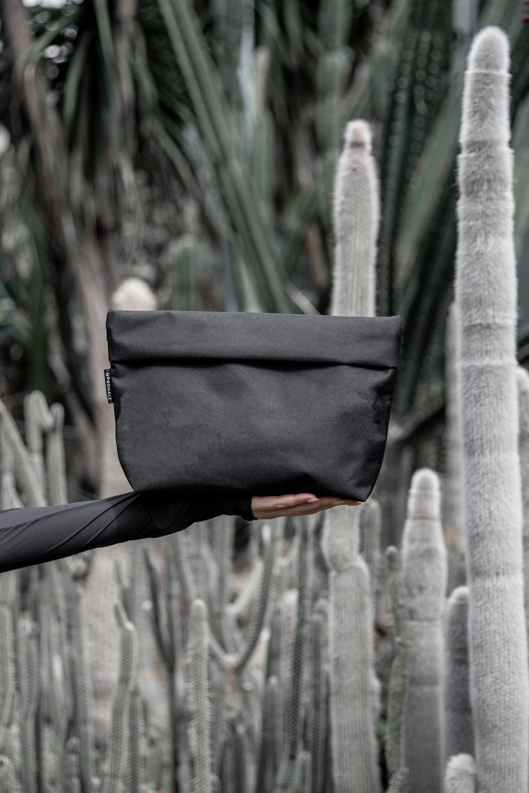 A hand holding a black Toiletry bag, perfect for travel enthusiasts. Water-resistant with YKK® zippers. Practical and lightweight design. Available in two sizes: Small (1.6L) and Medium (4.2L). Handmade from high-quality polyester canvas.