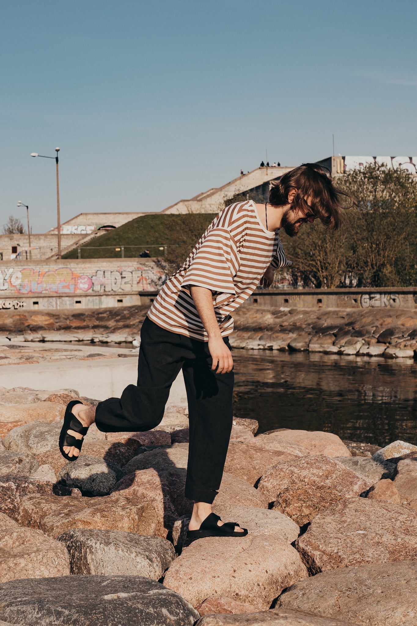 A man wearing Light Slip-On Leather Sandals walks on rocks by water.