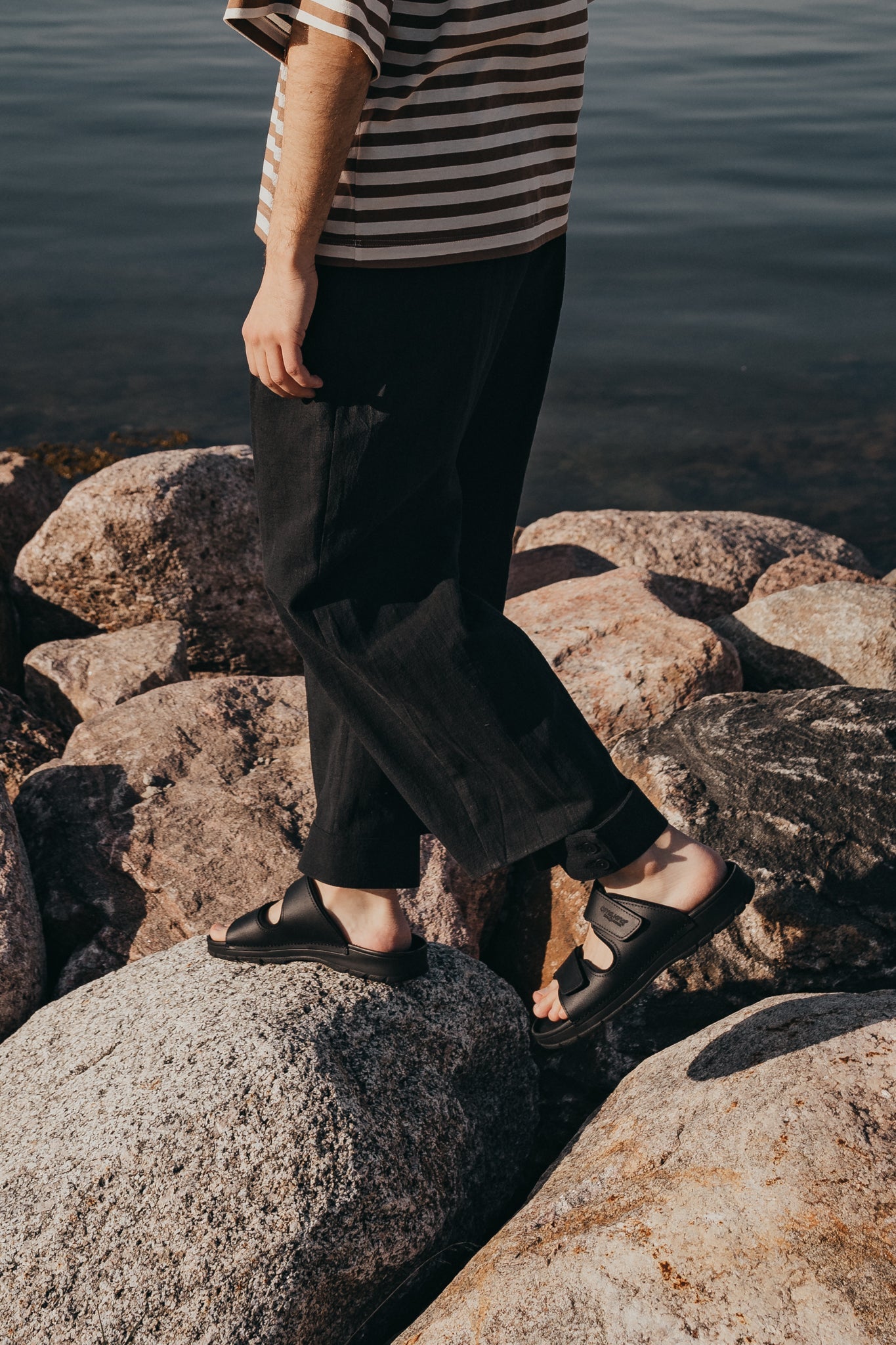 A person wearing Light Slip-On Leather Sandals stands on rocks near a lake.