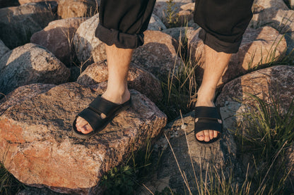 A person's feet in Slip-On Leather Sandals - Black on rocky terrain, showcasing comfort and style. Perfect for various settings, indoors, work, school, home, and outdoors. Designed and manufactured in Europe, these sandals combine quality materials and thoughtful construction for reliable and stylish footwear.