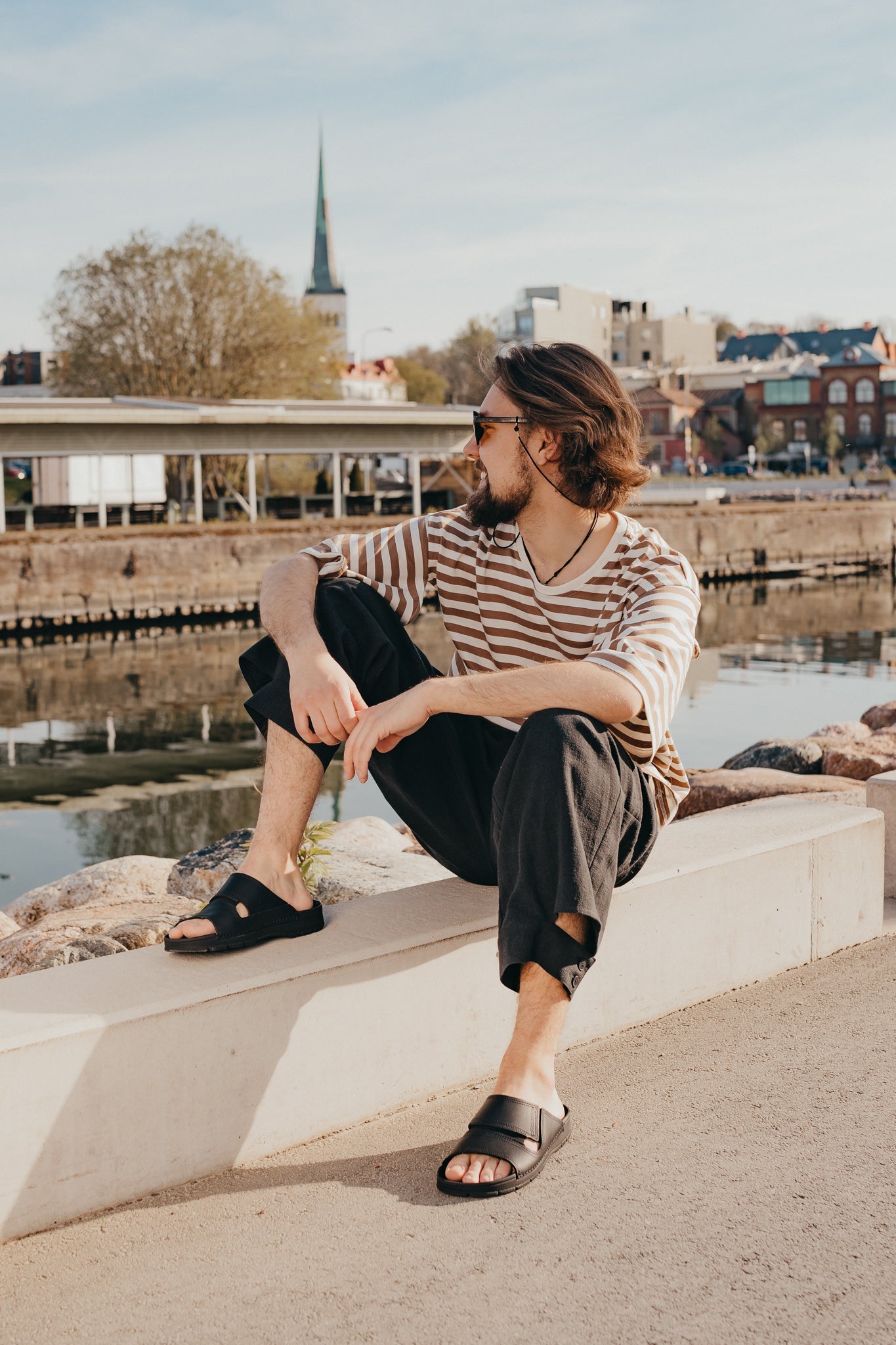 A man sitting on a ledge by water wearing Slip-On Leather Sandals - Black. Experience comfort and style with our versatile sandals for various settings.