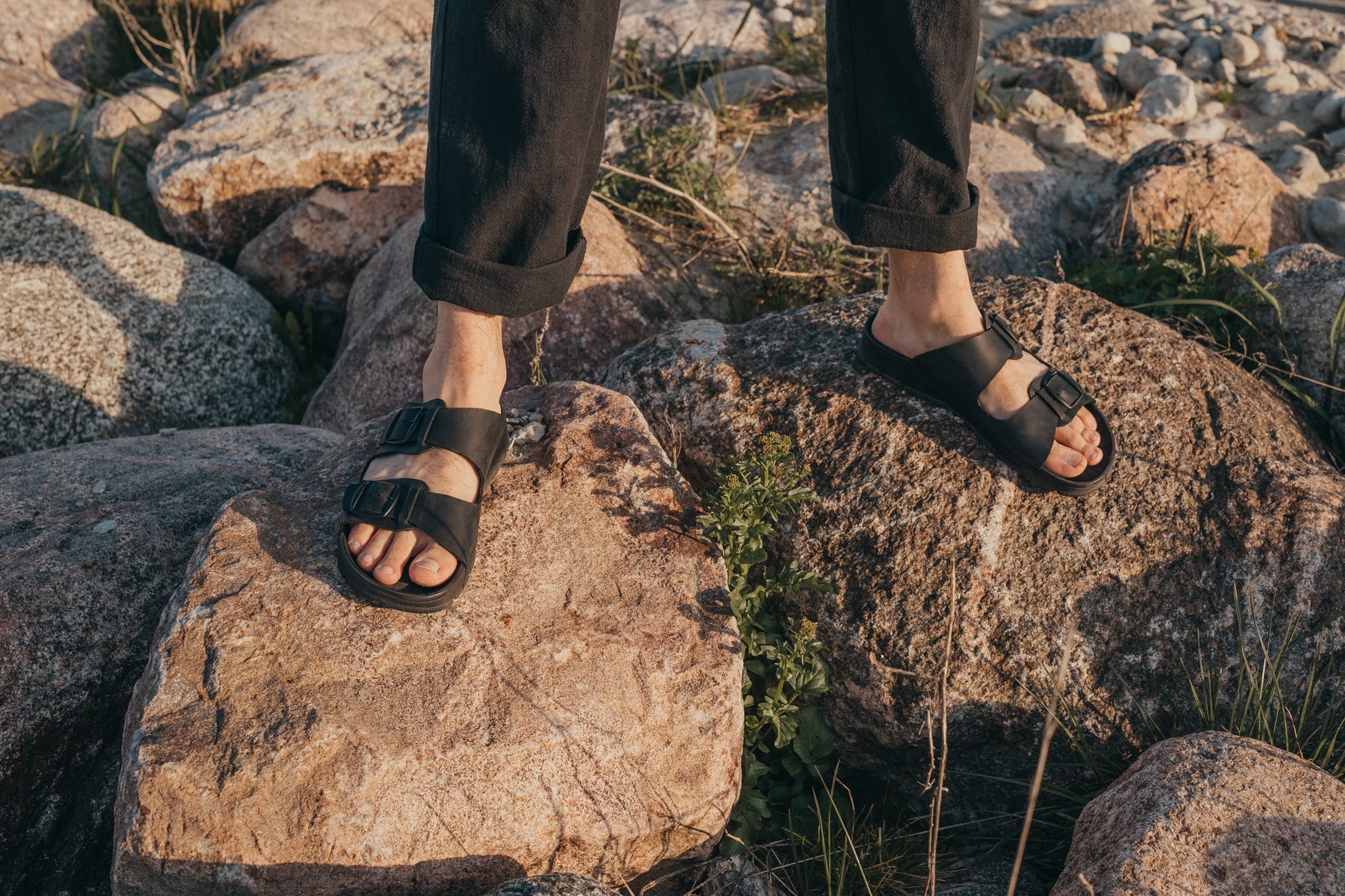 A person's feet in Slip-On Leather Sandals with Adjustable Buckle - Black, standing on rocks outdoors.